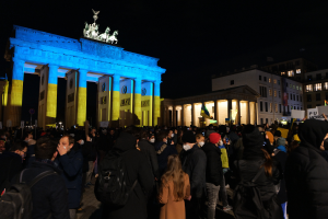 Eine Menschenmenge steht vor dem Brandenburger Tor in Berlin, Deutschland, viele tragen Mützen und Taschen, einige halten Schilder, mit den Säulen und Statuen des Tors im Hintergrund und Gebäuden mit Fenstern und einem dunklen Himmel.