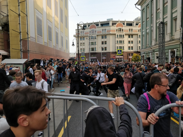 Eine große Menge steht hinter Metallabsperrungen auf einer Stadtstraße, einige halten Kameras, bei einer scheinbaren Demonstration.