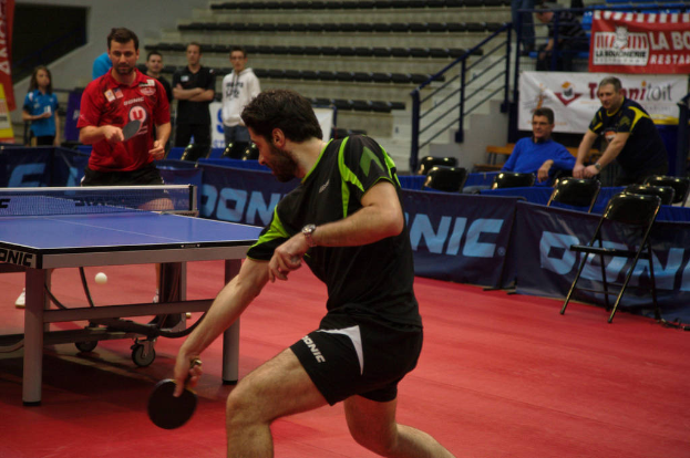 Eine Gruppe von Menschen in verschiedenen Farben, darunter Schwarz, Weiß und Blau, sitzt um einen Tisch mit einem Spiel in einem Raum mit Stühlen und einer Treppe im Hintergrund.