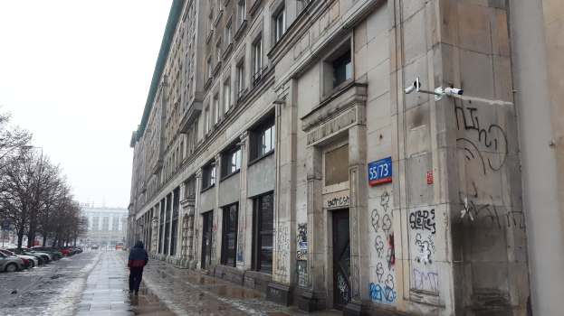 Eine Person, die auf einem Gehweg vor einem Graffiti-bedeckten Gebäude entlanggeht, mit parkenden Fahrzeugen, Bäumen und Gebäuden im Hintergrund.