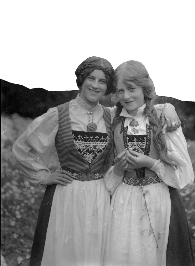 Zwei junge Frauen in traditioneller bayrischer Tracht, die weiße und schwarze Kleider mit aufwändiger Stickerei und Perlen tragen, posieren vor der Kamera und lächeln, vor einem Hintergrund aus üppigen grünen Pflanzen und einem hellen Himmel.