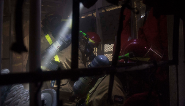 Eine Gruppe von Feuerwehrmännern in Helmen und Schutzausrütstung, einer mit einem Feuerlöscher, mit einem Metallgitter und einem Seil im Vordergrund und verschiedenen Gegenständen im Hintergrund.