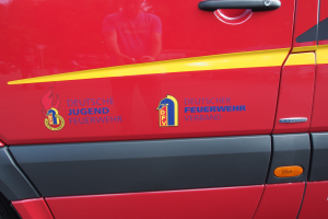 Ein roter Van mit einem gelben Streifen, einer Türklinke und einem Logo mit einem Schild, einem Löwen und einem Lorbeerkranz, sowie der Inschrift "Deutscher Jugendfeuerwehrverband".
