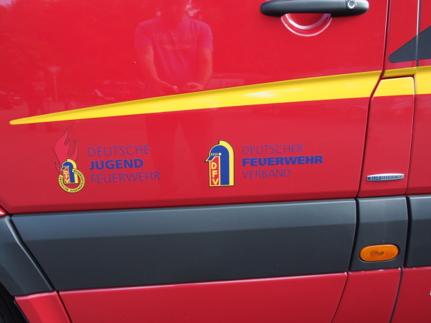 Ein roter Van mit einem gelben Streifen, einer Türklinke und einem Logo mit einem Schild, einem Löwen und einem Lorbeerkranz, sowie der Inschrift "Deutscher Jugendfeuerwehrverband".