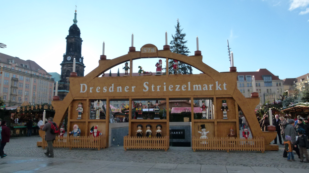 Ein geschäftiger Weihnachtsmarkt in Dresden, Deutschland, mit Menschen, die spazieren gehen, einem Torbogen, Bäumen, Gebäuden und bewölktem Himmel.