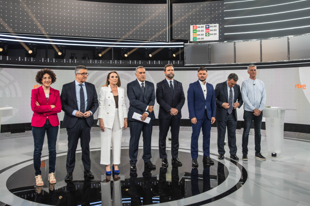 Eine Gruppe von Menschen steht vor einem Fernseher, flankiert von zwei Tischen mit verschiedenen Gegenständen, in einem Studio mit einer beleuchteten Wand und einem Bildschirm, bei einer Livesendung der spanischen Fernsehshow 'Rive'.