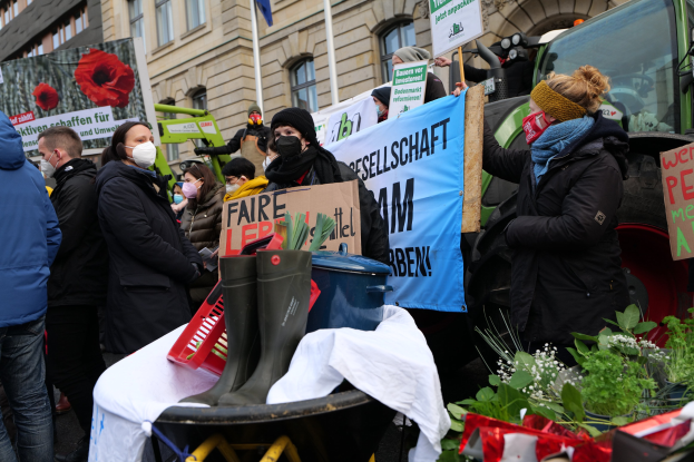 Eine Gruppe von Menschen mit Masken und Protestschildern vor einem Lastwagen, mit einem Tisch voller Gegenstände, Topfpflanzen und einer Flagge im Hintergrund.
