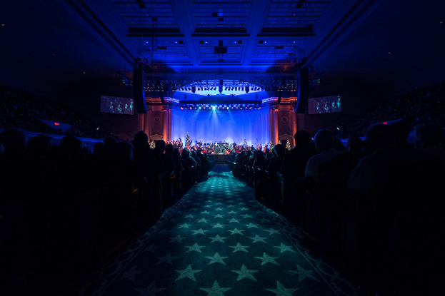 Ein großer Auditorium mit sitzenden Menschen, eine Bühne mit Musikern, die Instrumente spielen, und eine Decke mit Lichtern und Fernsehern, was auf einen Konzertsaal für die Nationale Konferenz 2019 hindeutet.