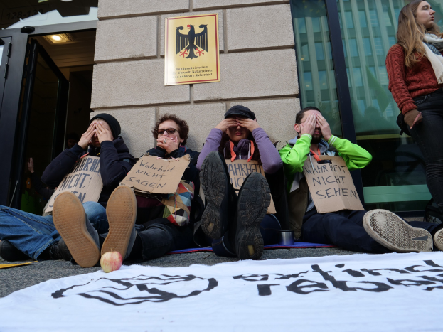 Menschen sitzen vor einem Gebäude auf dem Boden und halten Schilder, mit einerBanner auf der Straße und einer Frau rechts daneben, bei einer Protestaktion gegen eine Entscheidung der deutschen Regierung.