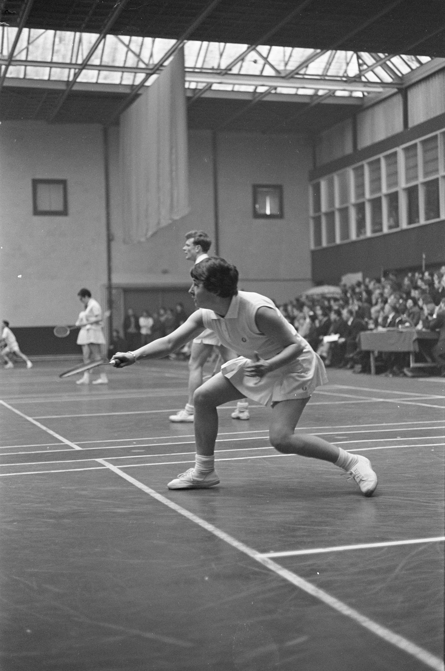 Schwarzes und weißes Foto von zwei Frauen, die Badminton auf einem Court spielen, jede hält einen Schläger, mit Zuschauern rechts und einer fensterumrandeten Wand im Hintergrund.