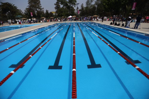 Großer Schwimmbad mit Bahnentrennungen, umgeben von Menschen mit Taschen und Gegenständen, mit Zelten, Transparenten, Stangen, einem Schild und einem bewölkten Himmel im Hintergrund.