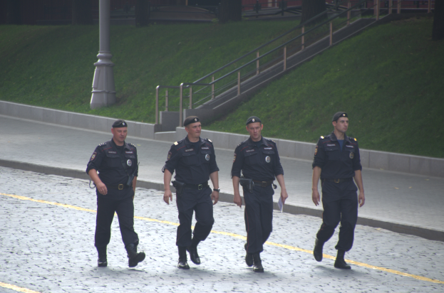 Vier Polizisten in Uniform gehen eine Straße entlang und halten Papiere in der Hand, mit einem Pfahl, Treppe, Geländer, Gras, Bäumen und einem Gebäude im Hintergrund.