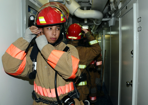 Feuerwehrleute in Uniform in einem Raum während einer Übung, mit Rohren und Equipment im Hintergrund.