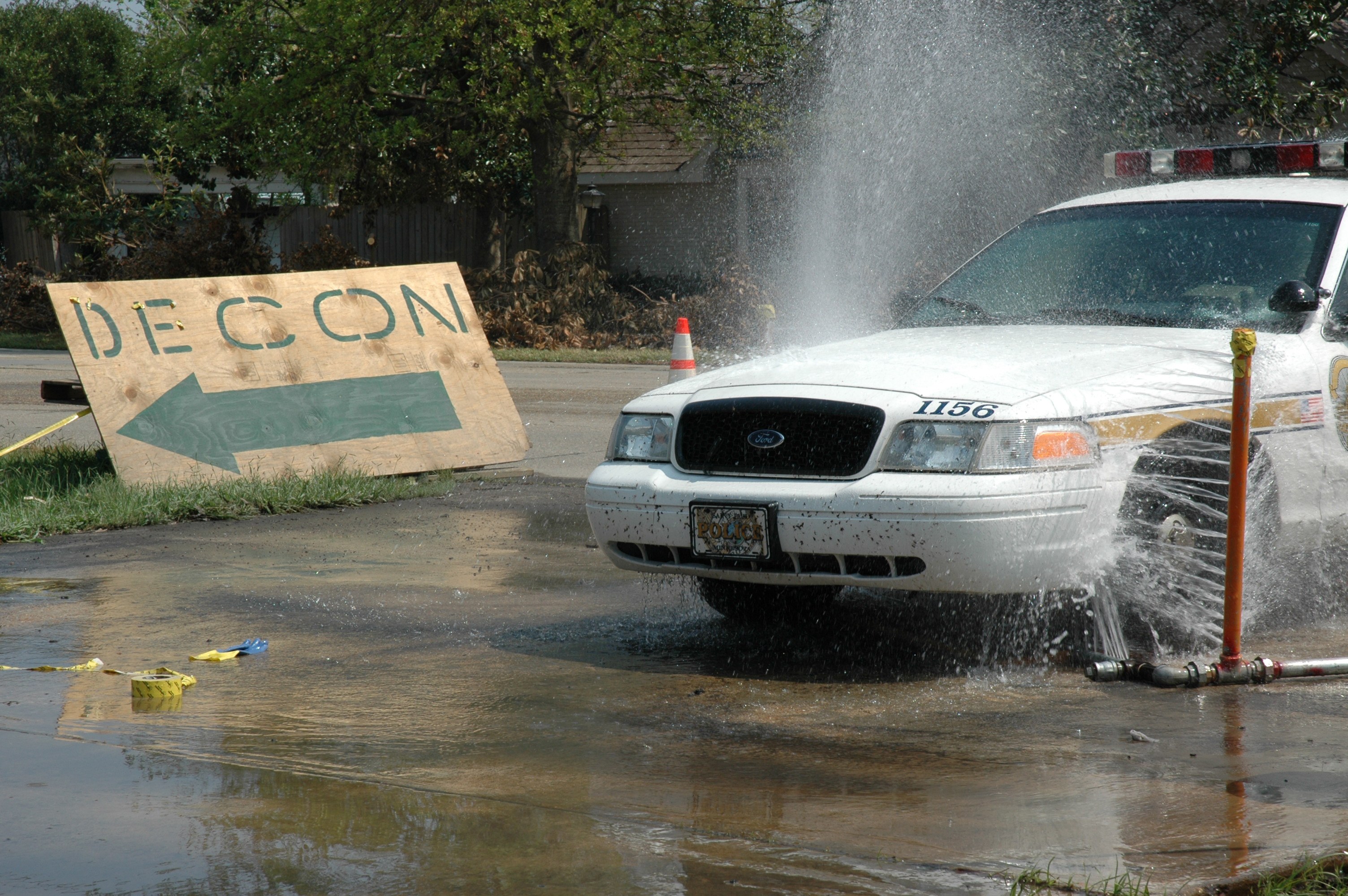 Ein Polizeiwagen fährt durch eine Pfütze, mit einem Schild, einer Verkehrskegel und anderen Gegenständen am Boden, vor Bäumen, Gebäuden und einem klaren Himmel.