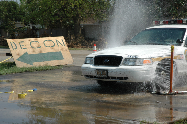 Ein Polizeiwagen fährt durch eine Pfütze, mit einem Schild, einer Verkehrskegel und anderen Gegenständen am Boden, vor Bäumen, Gebäuden und einem klaren Himmel.