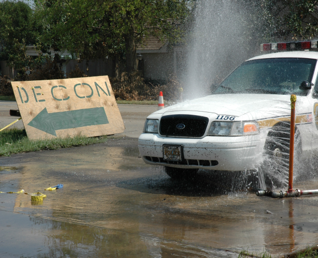 Ein Polizeiwagen fährt durch eine Pfütze, mit einem Schild, einer Verkehrskegel und anderen Gegenständen am Boden, vor Bäumen, Gebäuden und einem klaren Himmel.