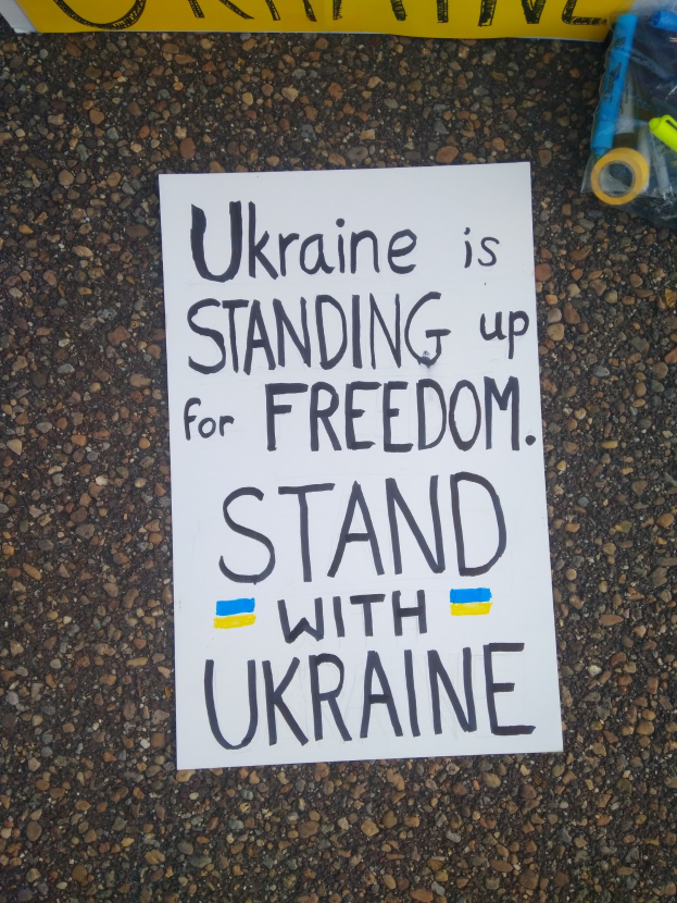 Ein weißes Blatt Papier mit dem Text "Ukraine steht für Freiheit, steht mit Ukraine" darauf, vor einem gelben Banner platziert, mit einer Plastikhülle mit verschiedenen Gegenständen rechts daneben.