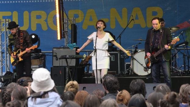 Eine Frau in einem weißen Kleid singt auf der Bühne bei einem Konzert, flankiert von zwei Gitarristen, mit einer Menge im Vordergrund und Lautsprechern im Hintergrund.
