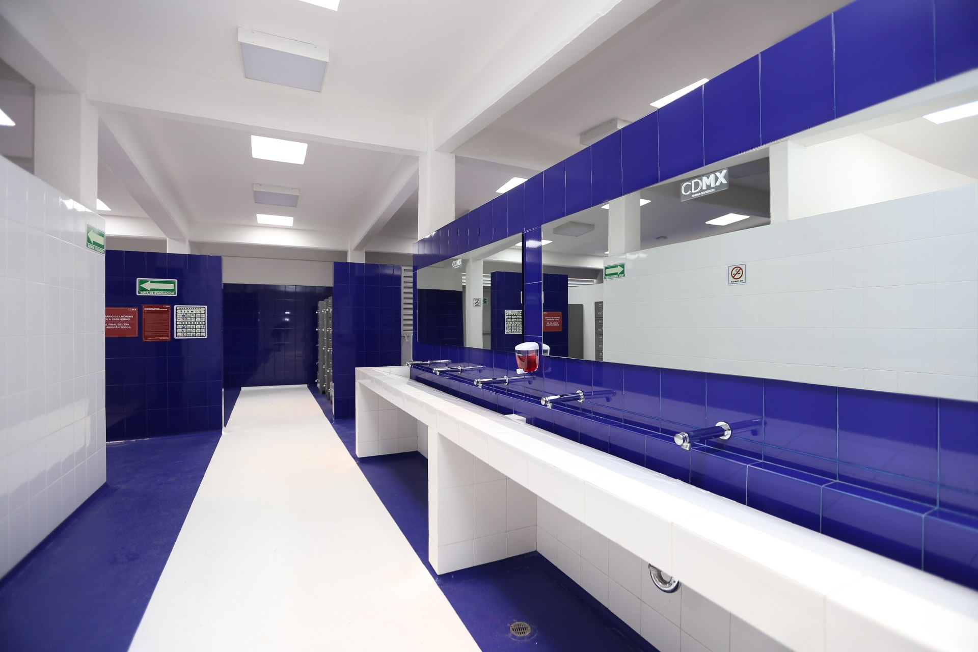 Öffentliche Toilette mit blauen Kacheln, weißen Tresen, Waschbecken mit Wasserhähnen, einer Plattform mit einer Flasche und Gegenständen, Informationsschildern an den Wänden, Deckenleuchten und Säulen.