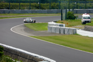 Ein Rennauto fährt auf einer Rennstrecke mit einem Polizeiwagen im Hintergrund, umgeben von Gras, Pflanzen, Bäumen und Absperrungen, mit einem Fahrzeug auf der rechten Seite und Text unten.