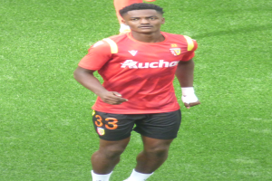 Emmanuel Adebayor, der ein rotes Hemd und schwarze Shorts trägt, läuft auf einem Fußballfeld mit zwei Personen im Hintergrund.