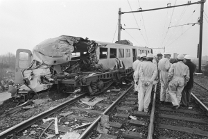 Ein Schwarz-Weiß-Bild einer Zugkollision mit Menschen in Helmen drumherum, Strommasten, Bäumen, Gebäuden und einem klaren Himmel im Hintergrund.