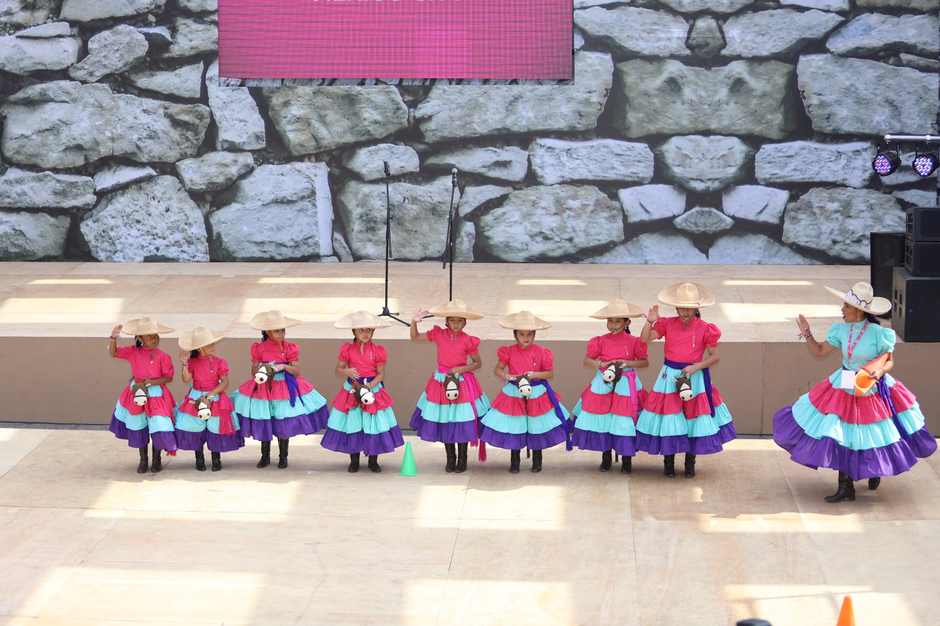 Eine Gruppe junger Mädchen in bunten Kleidern und Hüten tanzt auf einer Bühne mit Verkehrskegeln, Mikrofonen, Lautsprechern, Lichtern und einem Bildschirm mit Text im Hintergrund, während Menschen unten zusehen.