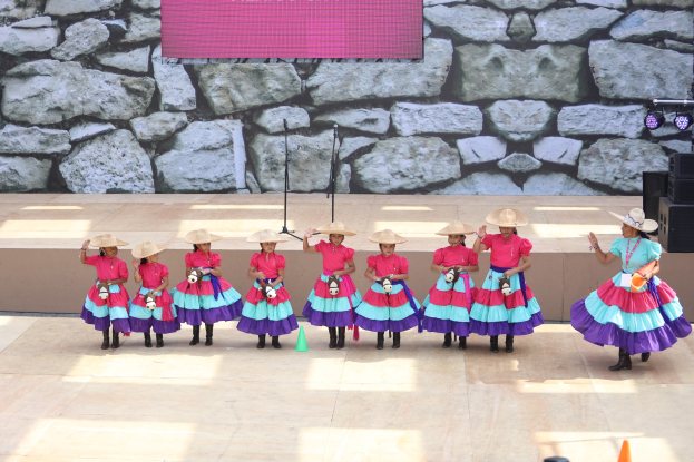 Eine Gruppe junger Mädchen in bunten Kleidern und Hüten tanzt auf einer Bühne mit Verkehrskegeln, Mikrofonen, Lautsprechern, Lichtern und einem Bildschirm mit Text im Hintergrund, während Menschen unten zusehen.