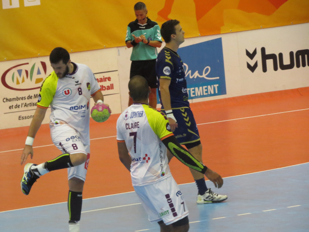 Mannergruppe spielt Handball auf einem Feld, einer hält den Ball, mit Bannern im Hintergrund.