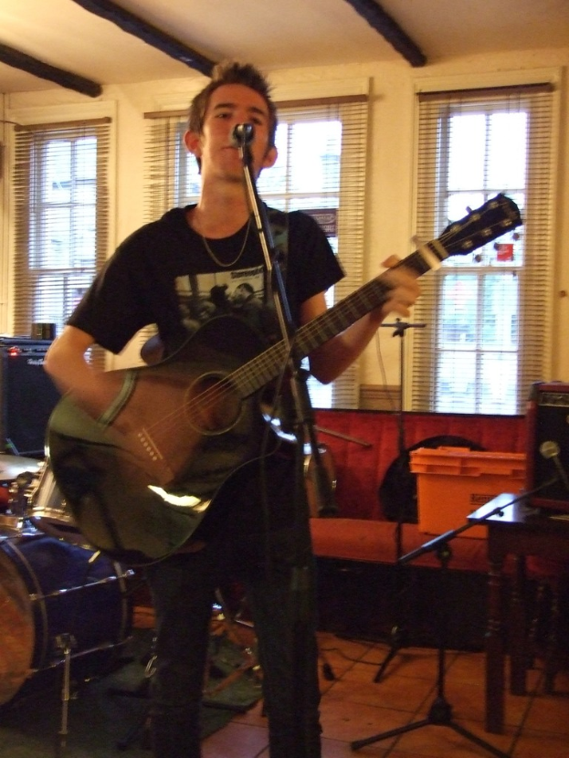 Ein Mann in schwarzem T-Shirt und Hose steht mit einer Gitarre und einem Mikrofon auf einem Ständer, mit einer blauen Trommel in der linken Ecke, einer Couch mit Gegenständen darauf und durchsichtigen Vorhängen im Hintergrund.