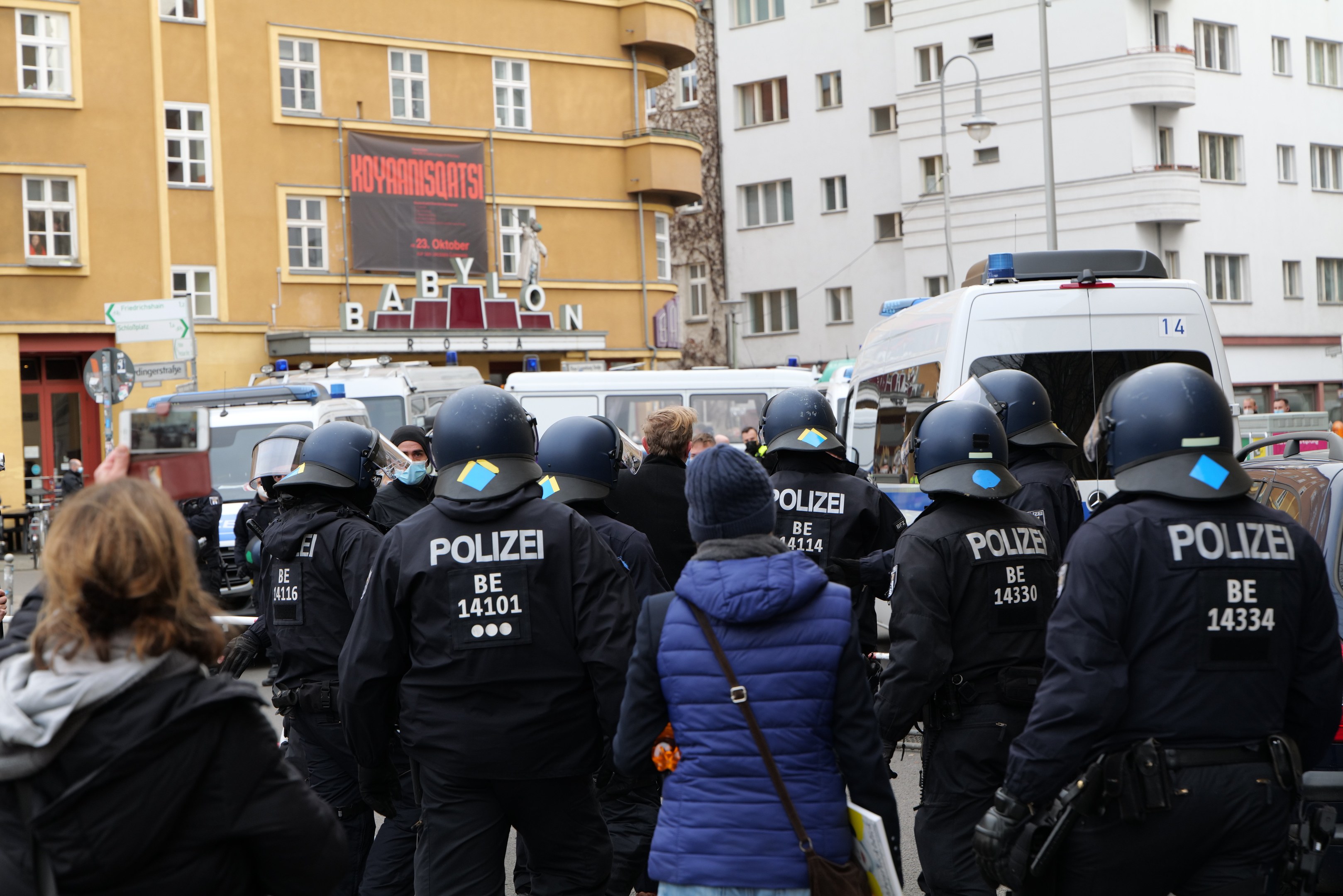 Polizeibeamte in Uniform vor einer Menge von Demonstranten, einige mit Helmen und Jacken, mit Fahrzeugen, Gebäuden und Laternen im Hintergrund während einer Demonstration in Berlin, Deutschland.