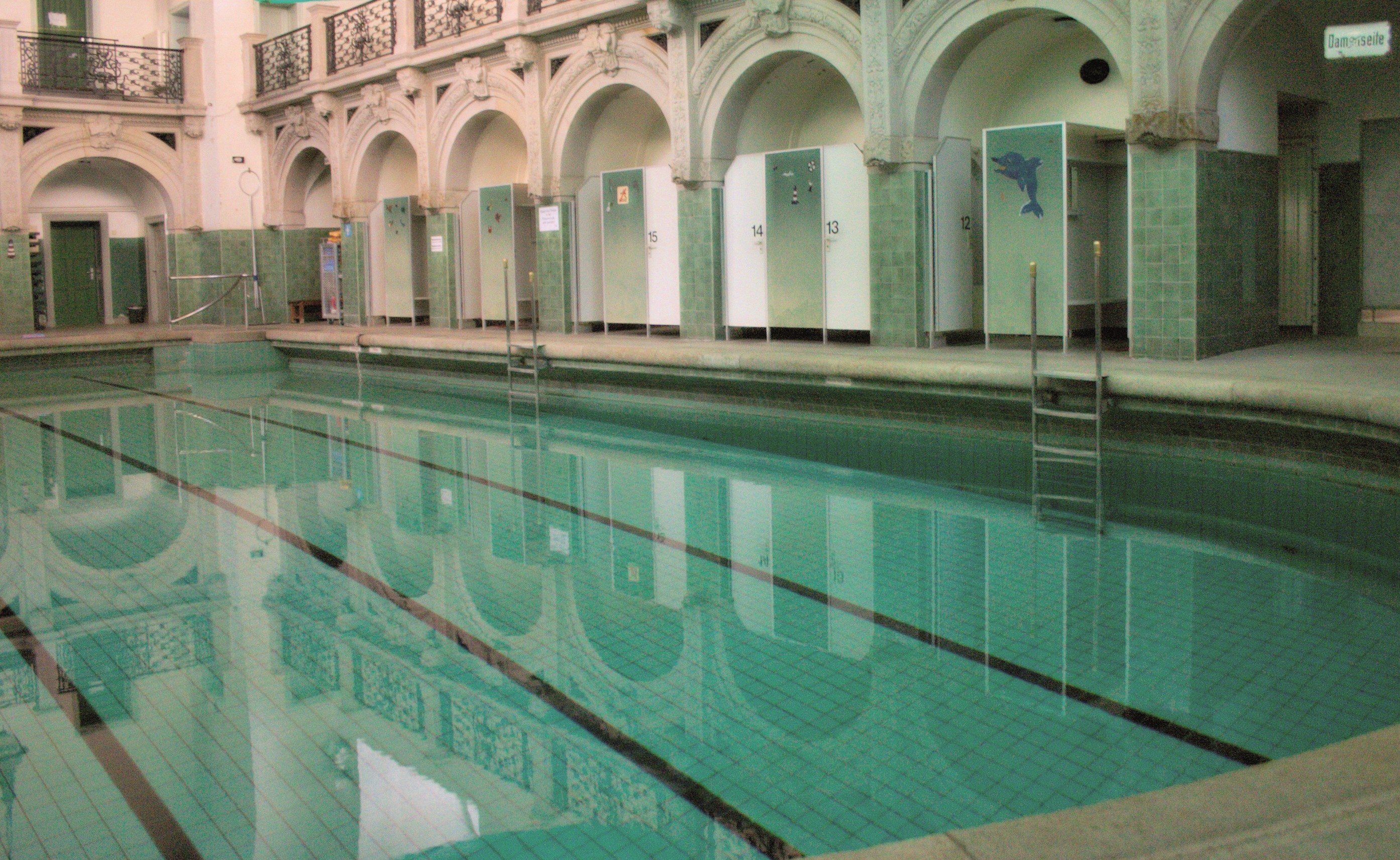 Innenbad mit klarem Wasser, umgeben von gefliestem Boden, in einem großen gewölbten Gebäude mit Säulen, Geländern, Türen und Texttafeln.