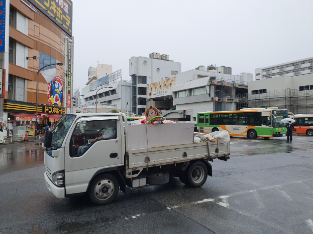 Ein weißer Lastwagen fährt eine Straße mit hohen Gebäuden entlang, mit Menschen darin, die Schirme halten, beleuchtet von Straßenlaternen und Schildern, unter einem sichtbaren Himmel.