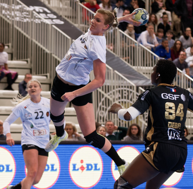 Eine Gruppe von Frauen, die Handball auf einem Platz spielen, mit einem Ball in der Luft, während Zuschauer im Hintergrund zuschauen.