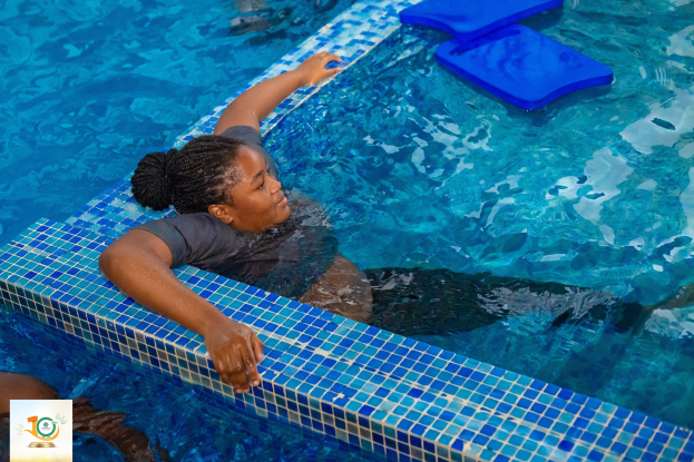 Eine Frau in einem T-Shirt mit ausgestreckten Armen in einem Schwimmbecken, umgeben von blauen Gegenständen, während einer Physiotherapie-Sitzung.