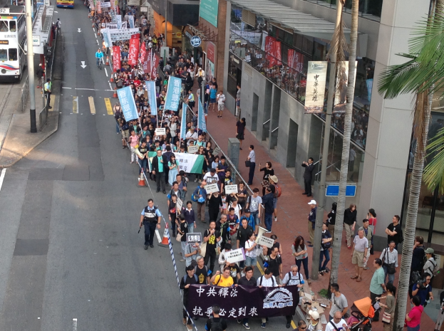 Eine große Gruppe von Menschen marschiert auf einer Straße in Hong Kong, wobei sie Protestschilder halten, mit Bäumen und gläsernen Gebäuden auf der rechten Seite und Fahrzeugen auf der linken Seite.