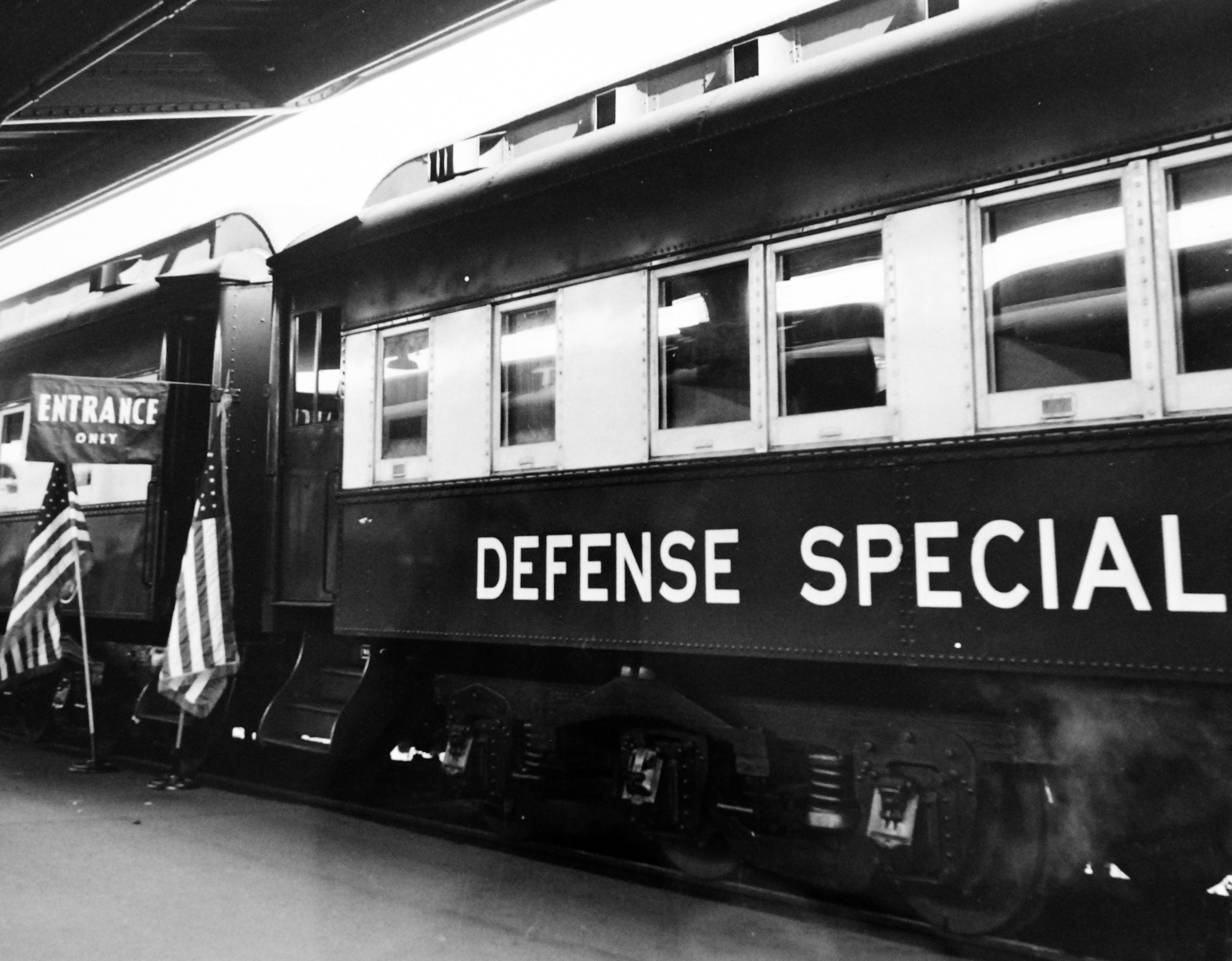 Schwarzes und weißes Foto eines Zuges mit der Aufschrift "Defense Special" auf den Eisenbahnschienen mit zwei Fahnen links und einer Decke oben.