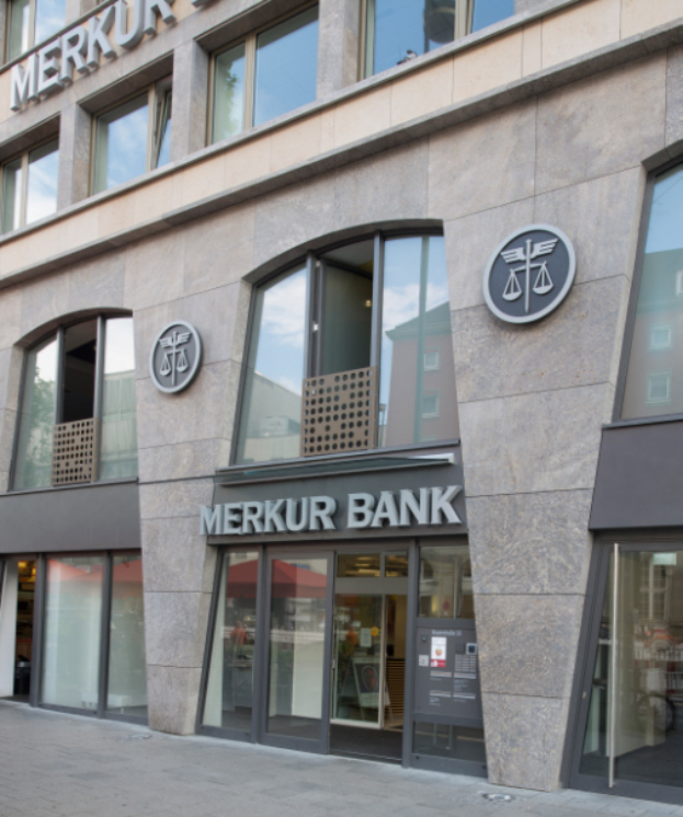 Ein modernes Gebäude mit Glaswänden und -türen, auf dem die Aufschrift "Merkur Bank" zu sehen ist, neben einer Straße stehend.