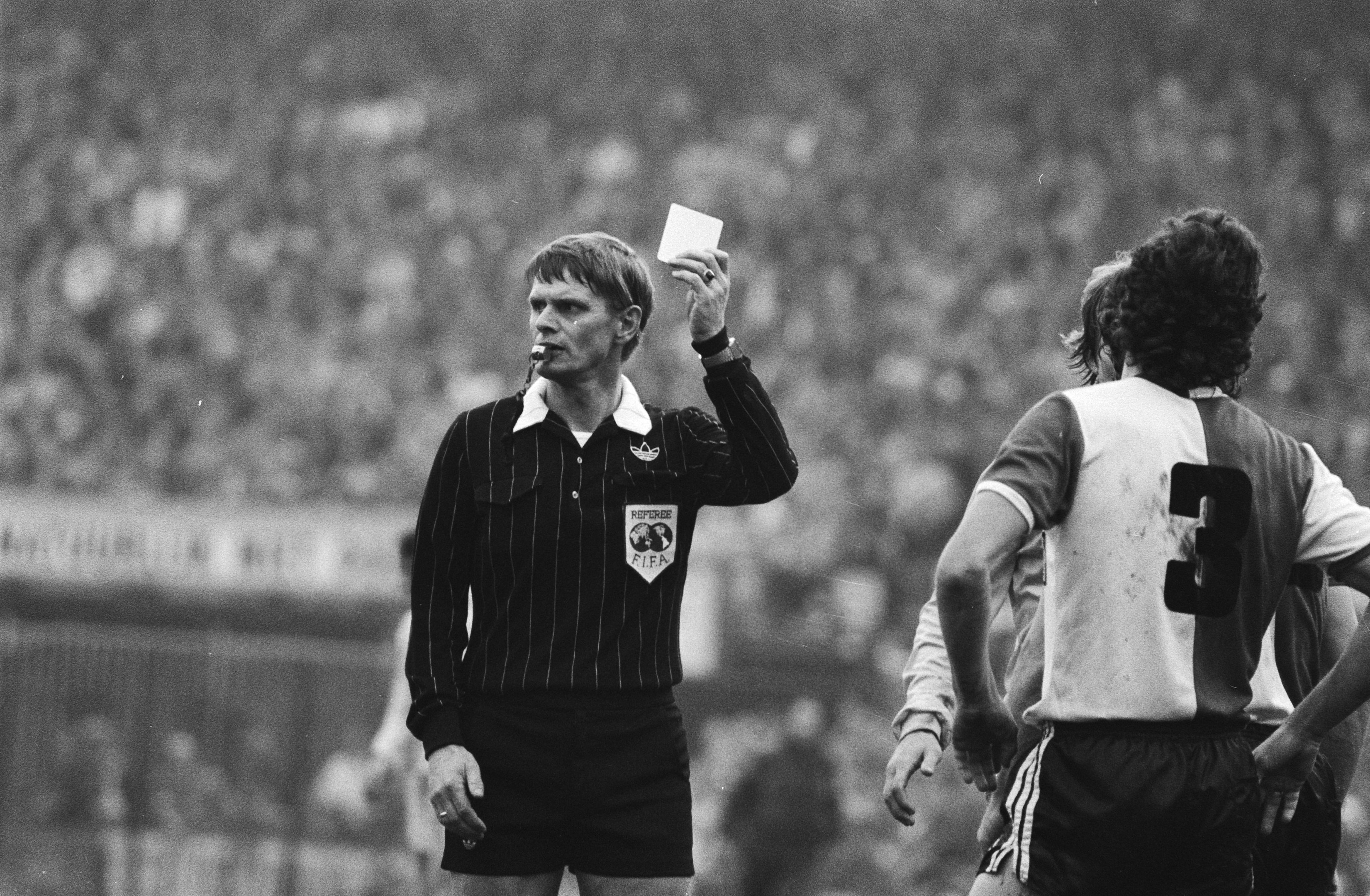 Schwarzes und weißes Bild eines Schiedsrichters, der eine rote Karte vor einer Menge in Fußballtrikots hält, mit einem Zaun im Hintergrund.