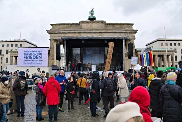 Eine Menschenmenge steht vor einem Gebäude mit einer Bühne, Lautsprechern und einem Bildschirm und hält Fahnen und Schilder, was auf eine Demonstration in Berlin hindeutet.