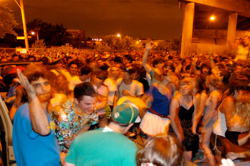 Menschen tanzen im Vordergrund eines Konzerts mit Lichtern, Pfählen, Bäumen und anderen Objekten im Hintergrund.