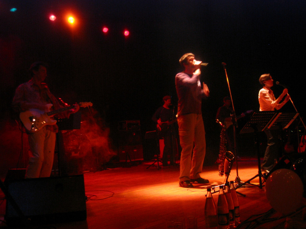 Zwei Menschen singen in ein Mikrofon auf einer Bühne mit drei Gitarristen und Wasserflaschen auf dem Podium.