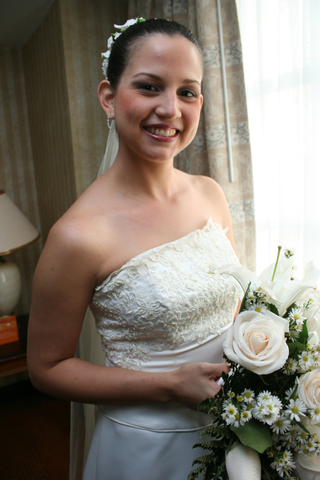 Eine Braut steht strahlend da und hält einen Strauß, mit einem Vorhang und einer Wand im Hintergrund und einer Lampe auf einem Tisch links.