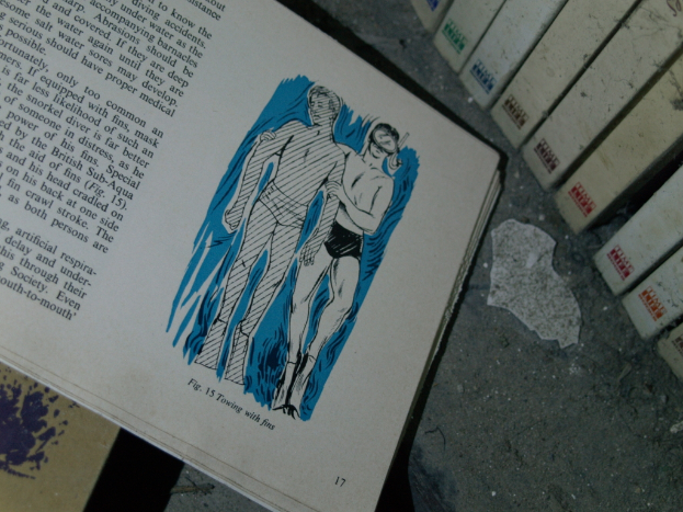 Ein aufgeschlagenes Buch mit Illustrationen einer Person in einem Tauchermasken-Taucher und einer anderen Person, mit anderen Büchern und Gegenständen im Hintergrund und Vordergrund.