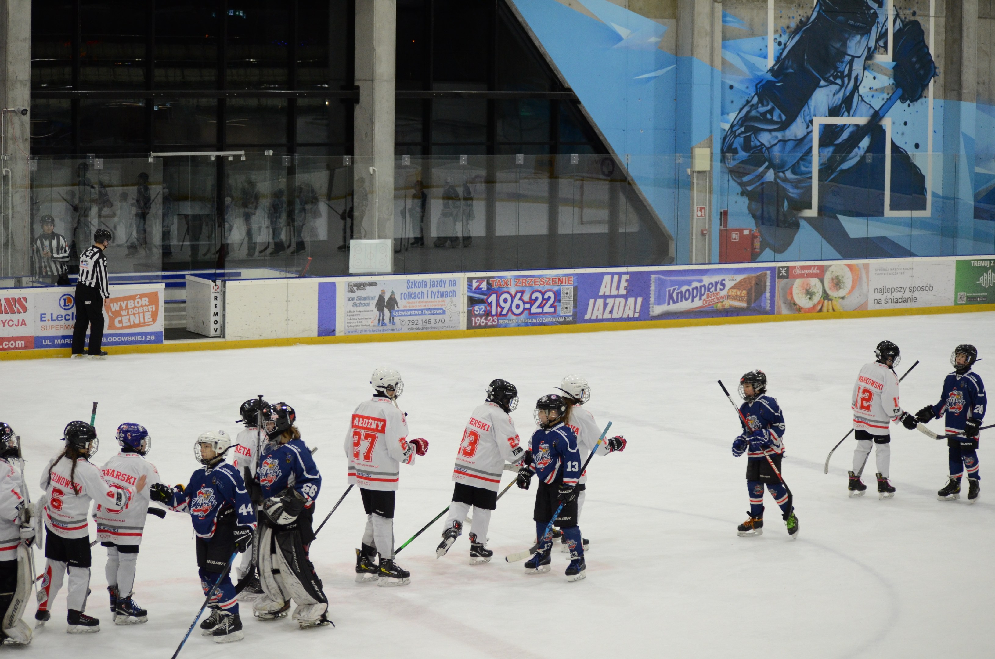 Junge Menschen in Helmen und Eishockeyausrüstung spielen Eis hockey auf einer Indoor-Eisbahn mit Bannern, Säulen und Glaswänden im Hintergrund.