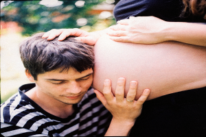 Ein Mann und eine Frau stehen nebeneinander, der Mann berührt sanft den Bauch der Frau und schaut auf ihr ungeborenes Kind.
