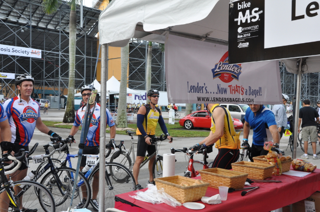 Eine Straßenszene mit Menschen, die in der Nähe von Fahrrädern stehen, ein Geschäft mit Gegenständen auf einem Tisch, Bäume, Zelte, an einem Schuppen befestigte Banner, Fahrzeuge und Gras.