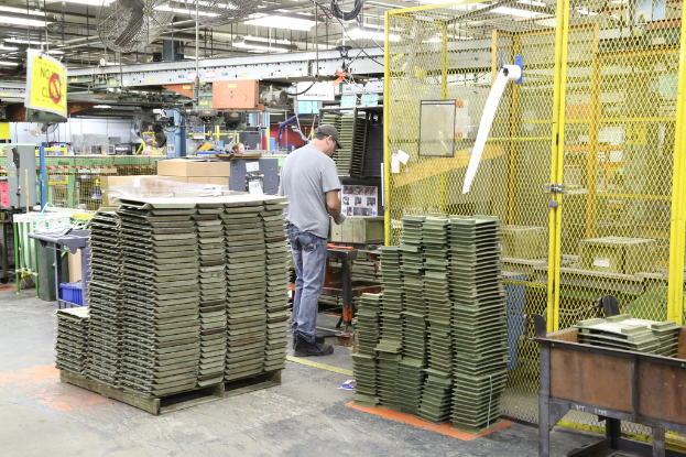 Ein Mann in einer Mütze und einem T-Shirt steht in einer Fabrik, arbeitet in der Nähe von Maschinen auf dem Boden und an der Decke, mit Lüftern und Lampen ebenfalls sichtbar.