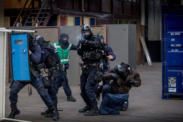 Polizeibeamte in Einsatzausrüstung stehen in einer Reihe, einige halten Waffen, mit einer Person auf dem Boden im Vordergrund und einem blauen Objekt mit Text auf der rechten Seite.