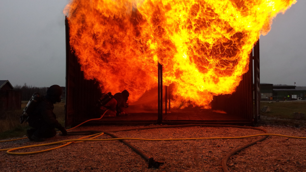 Feuerwehr löscht Gebäudebrand mit einer Leitung auf dem Boden, Schuppen und Bäume im Hintergrund unter einem klaren blauen Himmel.